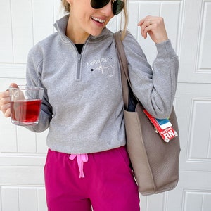 May include: A woman wearing a grey sweatshirt with the text "Kayla RN" and a stethoscope graphic, pink scrub pants, and a brown tote bag. She is holding a glass of red liquid and wearing sunglasses.
