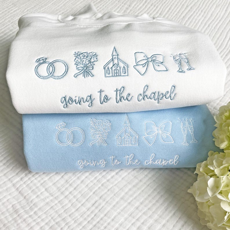 May include: Two sweatshirts, one white and one light blue, are stacked. Both have embroidered wedding-themed designs: rings, flowers, a church, a bow, and champagne glasses. The text "going to the chapel" is embroidered on each.