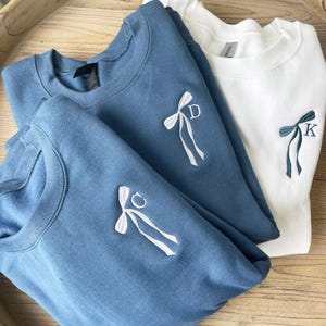 May include: Three sweatshirts with embroidered bows and initials. The top sweatshirt is white with a green bow and the initial "K". The middle sweatshirt is blue with a white bow and the initial "D". The bottom sweatshirt is blue with a white bow and the initial "C".
