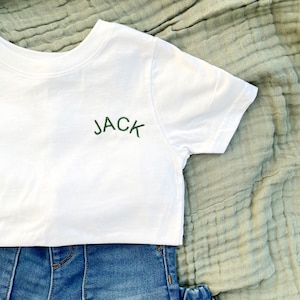 May include: White t-shirt with green embroidered text that says "JACK" on the front. The shirt is laying on a light green textured fabric.