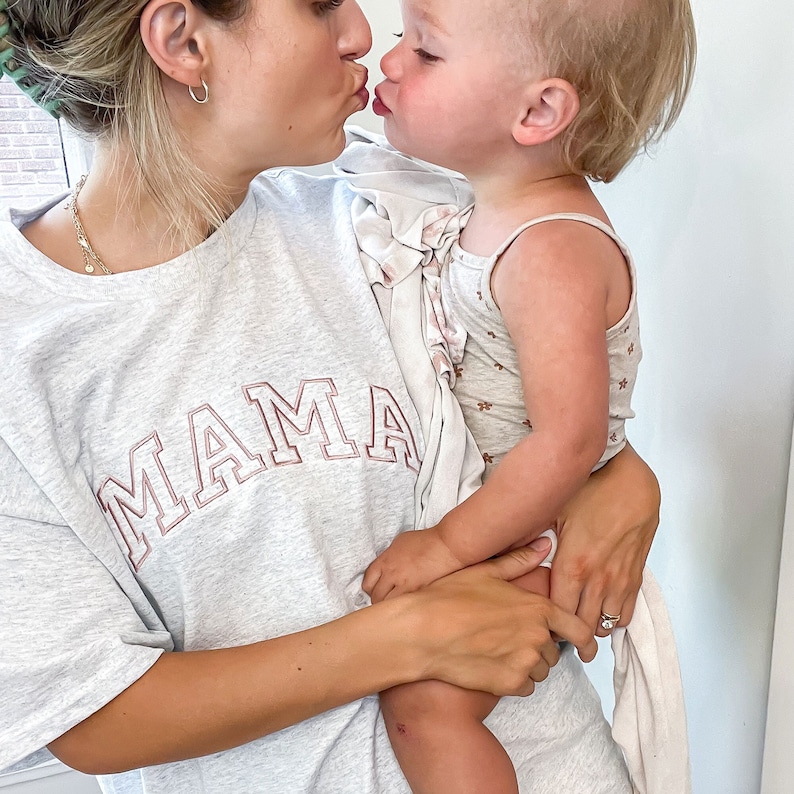 K&ouml;nnte beinhalten: Eine Frau tr&auml;gt ein graues T-Shirt mit dem Wort "MAMA" in Pink auf der Vorderseite und h&auml;lt ein kleines Kind in ihren Armen. Die Frau und das Kind k&uuml;ssen sich.