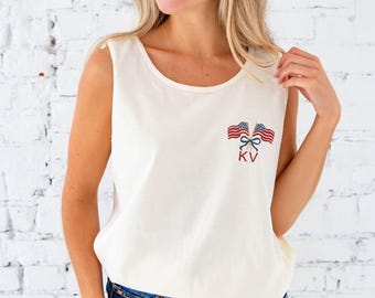May include: Cream-colored tank top with a scoop neck and a small embroidered design featuring two American flags tied with a bow and the initials "KV" in red. The top is sleeveless and made of a soft fabric.