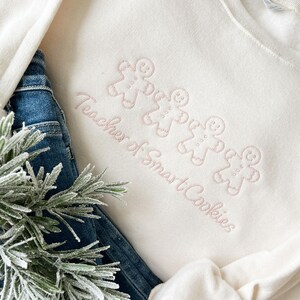 sweet cream crewneck sweatshirt with custom gingerbread men icons and "teacher of smart cookies" design embroidered in powder pink thread