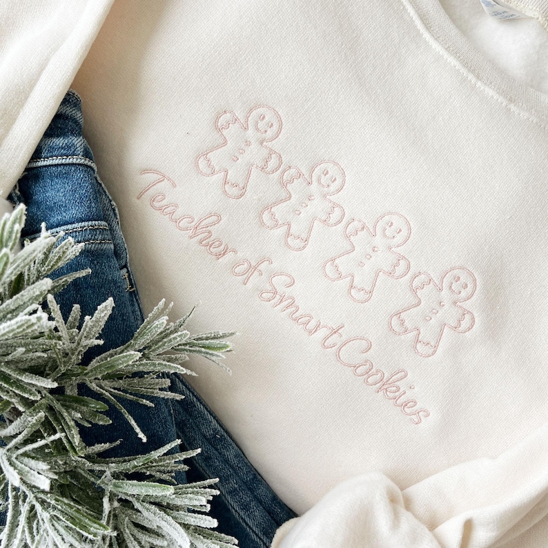 sweet cream crewneck sweatshirt with custom gingerbread men icons and "teacher of smart cookies" design embroidered in powder pink thread