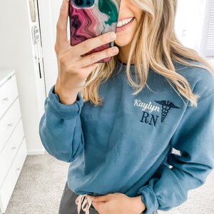 Op de afbeelding: Een blauw sweatshirt met de tekst "Kaylyn RN" en een geborduurd medisch symbool op de voorkant. Het sweatshirt wordt gedragen door een persoon met lang blond haar.