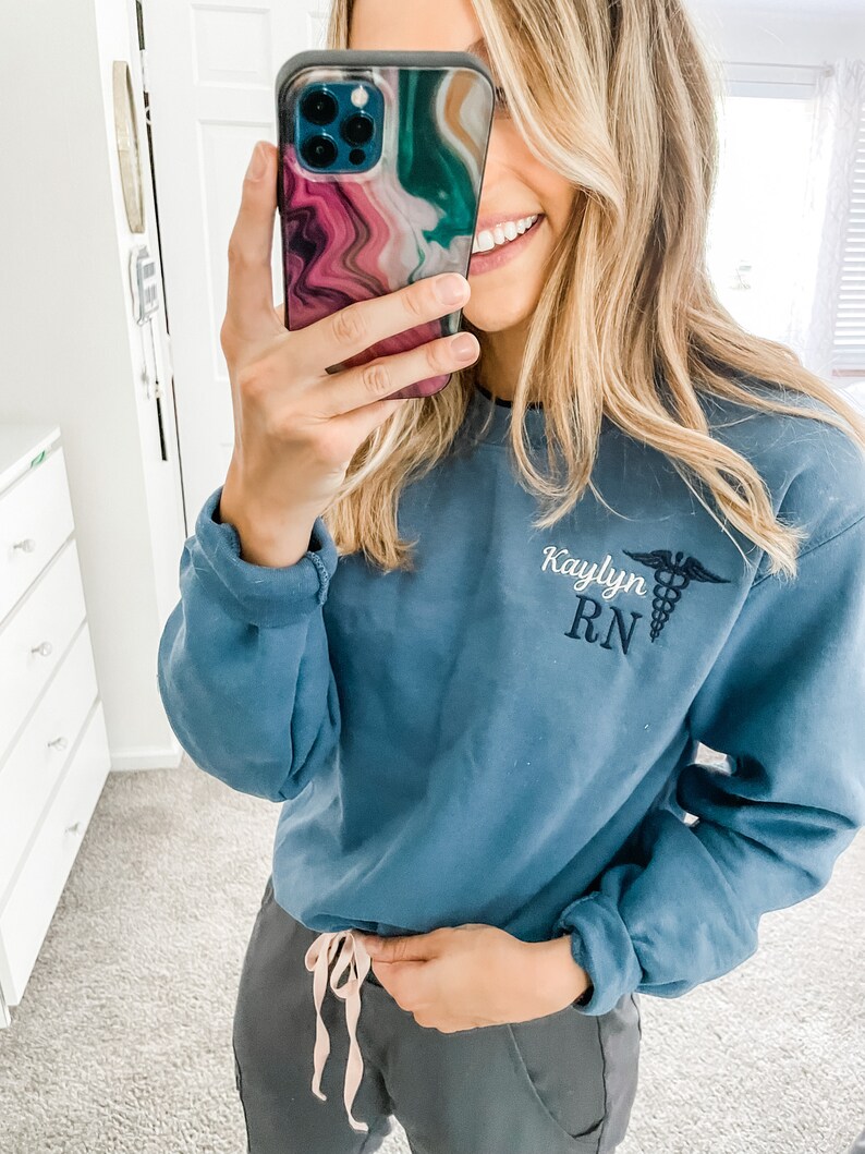 Op de afbeelding: Een blauw sweatshirt met de tekst "Kaylyn RN" en een geborduurd medisch symbool op de voorkant. Het sweatshirt wordt gedragen door een persoon met lang blond haar.