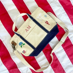 May include: Cream-colored canvas tote bag with navy blue trim and a cream-colored shoulder strap. The bag features embroidered designs of a bow, star, lobster, rose, and American flag. The initials "AM" are embroidered in red.