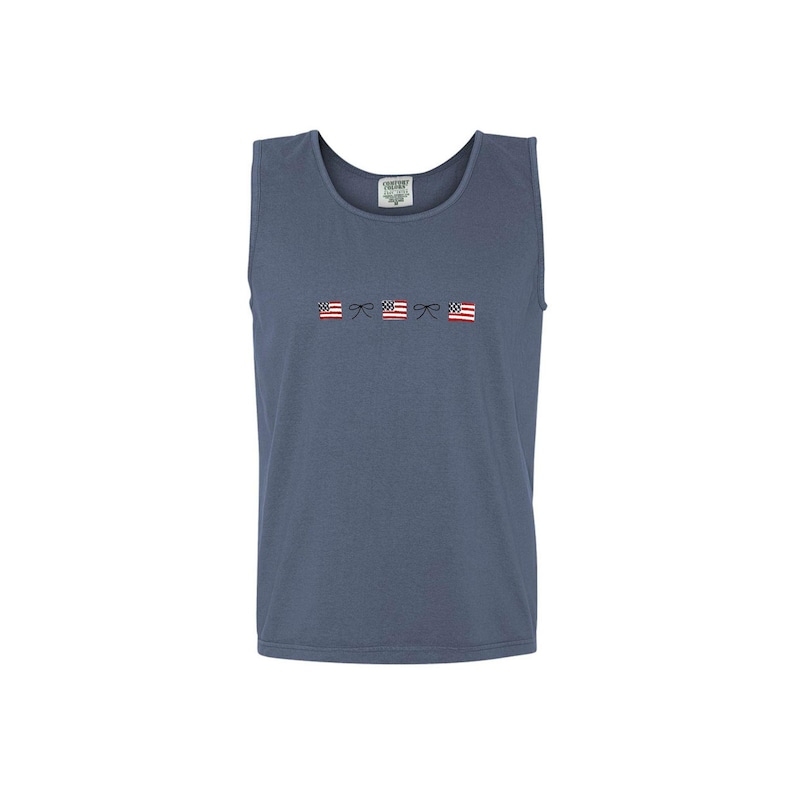 A blue jean comfort colors tank top with embroidered mini usa flags and dainty bow design across the chest in an alternating pattern