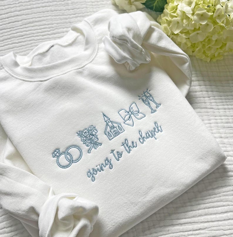 May include: White sweatshirt with light blue embroidered wedding-themed graphics, including rings, flowers, a church, a bow, and champagne glasses. The phrase "going to the chapel" is embroidered below the graphics.