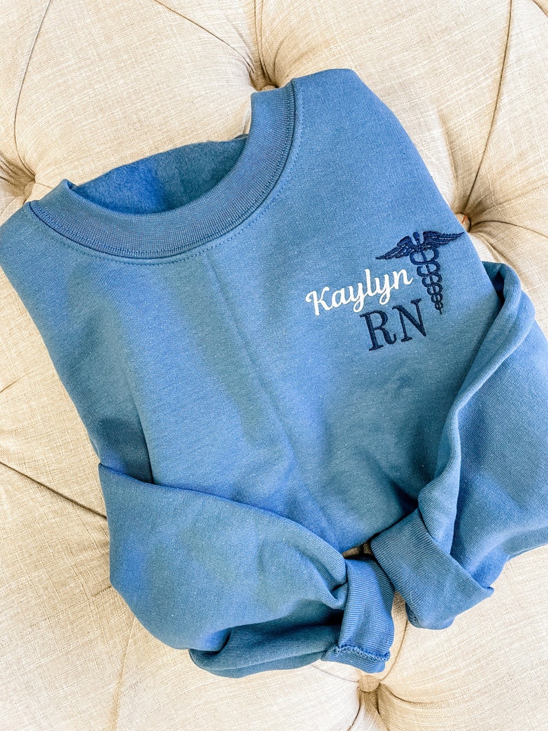 Op de afbeelding: Een blauw sweatshirt met de tekst "Kaylynn RN" en een geborduurd medisch symbool op de voorkant.