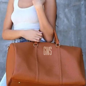 May include: A brown leather duffel bag with the initials "GWS" in gold lettering. The bag has two handles and a shoulder strap. The bag is a medium size and appears to be new.