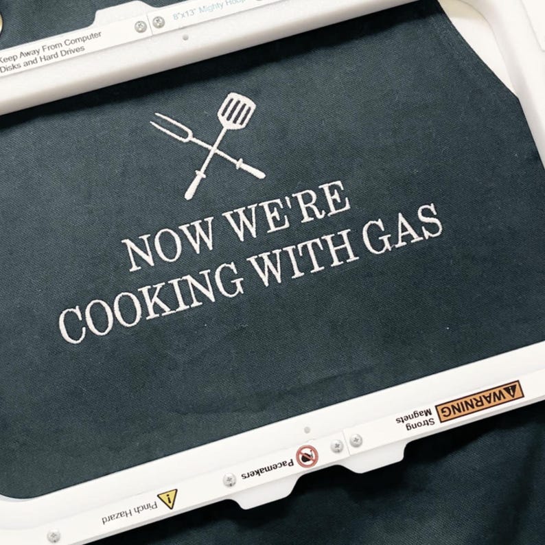close up of a charcoal apron with custom embroidered text reading: "now we're cooking with gas"