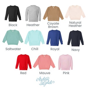 K&ouml;nnte beinhalten: Eine Auswahl von 12 Sweatshirts in verschiedenen Farben f&uuml;r Kleinkinder, darunter Schwarz, Heather Grey, Coyote Braun, Natur Heather, Salzwasser, Chill, Royalblau, Marine, Rot, Mauve und Pink. Die Sweatshirts sind in einem Raster auf wei&szlig;em Hintergrund angeordnet.