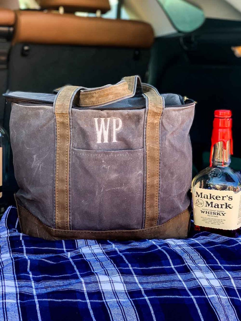 May include: A gray canvas tote bag with brown leather handles and a zipper closure. The bag has the initials "WP" embroidered on the front. A bottle of Maker's Mark whiskey is sitting next to the bag on a blue and white plaid blanket.