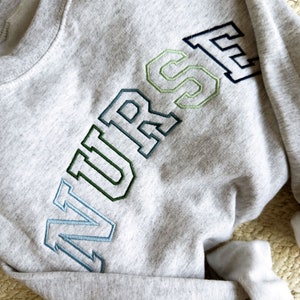 an ash crewneck sweatshirt with custom NURSE embroidered design in shades of blue and green