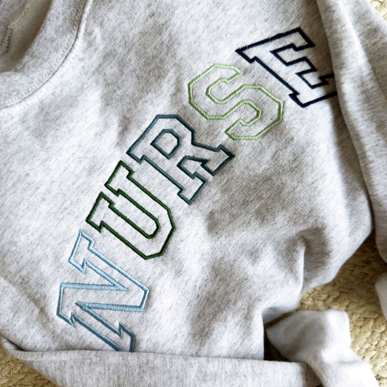 an ash crewneck sweatshirt with custom NURSE embroidered design in shades of blue and green