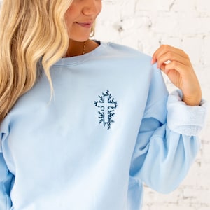 May include: A light blue sweatshirt with a dark blue embroidered cross design. The cross is surrounded by leaves.