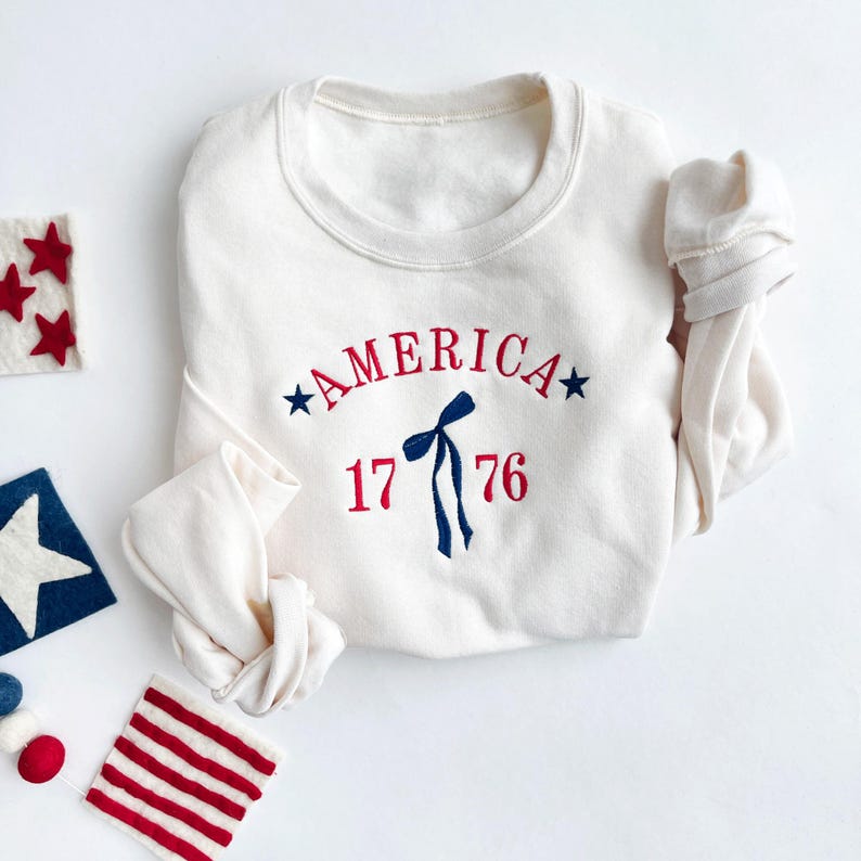 May include: Cream-colored sweatshirt with the word "AMERICA" arched across the chest in red, with blue stars on either side. Below, the number "1776" is embroidered in red. A blue bow and streamers are centered below the word "AMERICA".