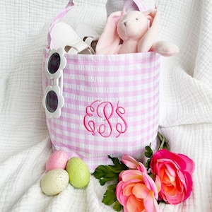 Peut inclure: Un sac cabas en gingham rose et blanc avec un monogramme rose "EBS" et un jouet de lapin rose à l'intérieur. Le sac est posé sur une surface blanche avec des œufs de Pâques roses et verts et des fleurs roses.