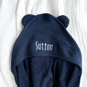 Może przedstawiać: Granatowy ręcznik kąpielowy dla niemowląt z kapturem i uszami misia. Imię "Sutton" jest wyhaftowane na kapturze jasnoniebieskim kolorem. Ręcznik wykonany jest z miękkiego materiału i leży na białej, teksturowanej powierzchni.