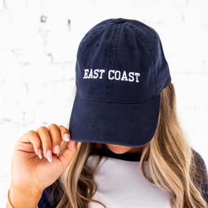 May include: Navy blue baseball cap with the white text "EAST COAST" embroidered on the front. The cap has a curved brim and a classic, adjustable design. The cap is made of a soft, durable material.