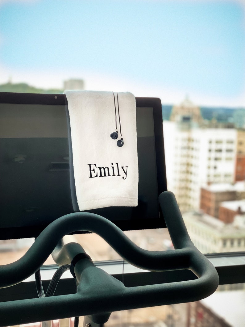 Puede incluir: Una toalla blanca con el nombre "Emily" bordado en ella est&aacute; colgada en una bicicleta est&aacute;tica con auriculares negros colgados sobre ella.