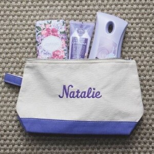 Pode incluir: Uma bolsa de lona branca com z&iacute;per, acabamento roxo e o nome "Natalie" bordado em roxo. A bolsa est&aacute; cheia de tr&ecirc;s artigos de higiene pessoal com aroma de lavanda.