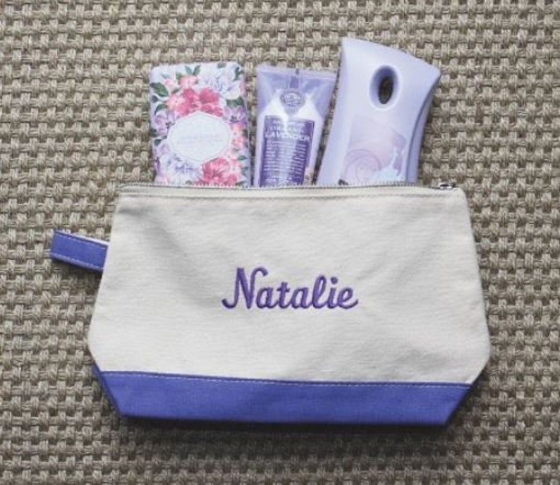 Pode incluir: Uma bolsa de lona branca com z&iacute;per, acabamento roxo e o nome "Natalie" bordado em roxo. A bolsa est&aacute; cheia de tr&ecirc;s artigos de higiene pessoal com aroma de lavanda.