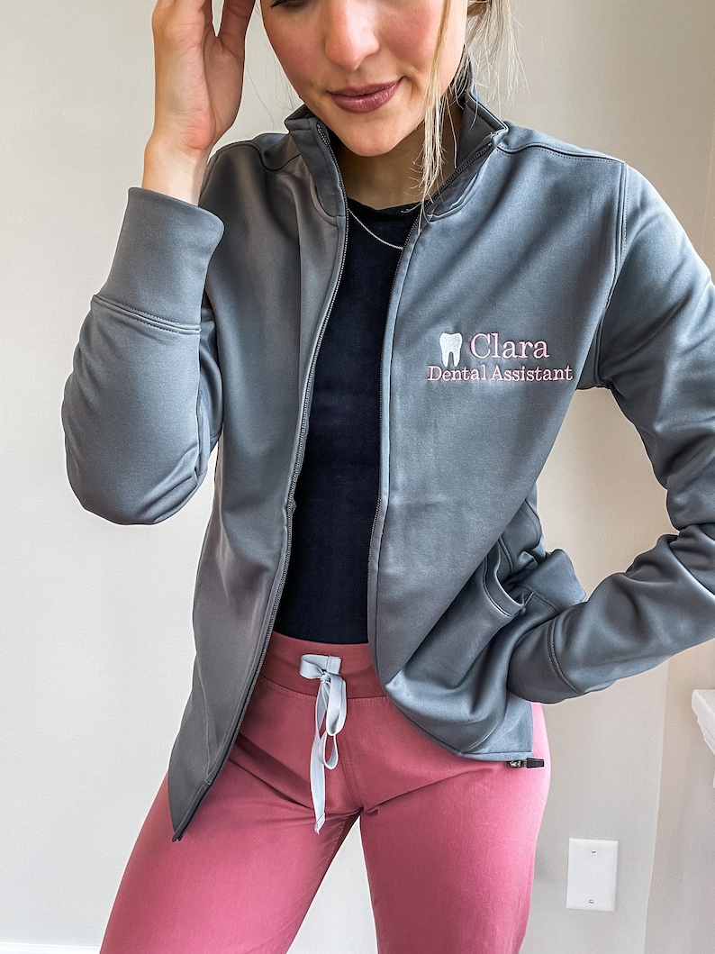 Puede incluir: Una chaqueta gris con cremallera con un logotipo de diente blanco y el texto "Clara Dental Assistant" bordado en la parte delantera. La chaqueta se lleva sobre una camisa negra y unos pantalones rosas.