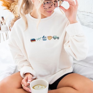May include: A white sweatshirt featuring a graphic design of a stack of books, a dragonfly, a gazebo, a cup of coffee, and a jeep. The sweatshirt is being worn by a person sitting on a white bed.