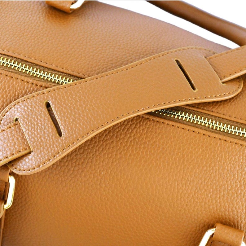 May include: Close-up of a tan leather bag with a shoulder strap. The bag features a gold zip and a textured surface. The shoulder strap is attached with a stitched seam and has a cut-out design.