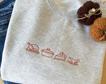 May include: A light gray crewneck sweatshirt featuring a minimalist red embroidered line art design of a roasted turkey, a covered casserole dish, a slice of pie, and a gravy boat. The garment is paired with blue denim jeans and two small pumpkins.