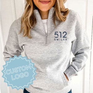 May include: A light gray pullover sweatshirt with a quarter-zip neckline and a front pocket. The sweatshirt features the text "512 SMILES" in navy blue. A light blue custom logo is in the lower left corner.