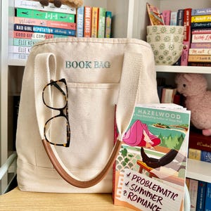 May include: A cream-colored canvas tote bag with the text "BOOK BAG" in light blue. The bag has a brown leather handle and a pair of tortoiseshell glasses hanging from it. A book titled "Problematic Summer Romance" is visible.