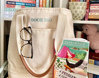 Könnte beinhalten: Eine cremefarbene Canvas-Tragetasche mit dem Text "BOOK BAG" in Hellblau. Die Tasche hat einen braunen Ledergriff und eine Brille mit Schildpattmuster. Ein Buch mit dem Titel "Problematic Summer Romance" ist sichtbar.