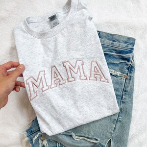 K&ouml;nnte beinhalten: Ein graues T-Shirt mit dem Wort "MAMA" in rosa auf der Vorderseite gestickt. Das Shirt ist gefaltet und auf einer blauen Jeanshose gelegt.