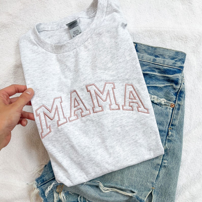 K&ouml;nnte beinhalten: Ein graues T-Shirt mit dem Wort "MAMA" in rosa auf der Vorderseite gestickt. Das Shirt ist gefaltet und auf einer blauen Jeanshose gelegt.
