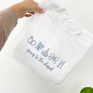 May include: White sweatshirt with light blue embroidered wedding-themed icons, including rings, flowers, a church, a bow, and champagne glasses. The phrase "going to the chapel" is embroidered below the icons.