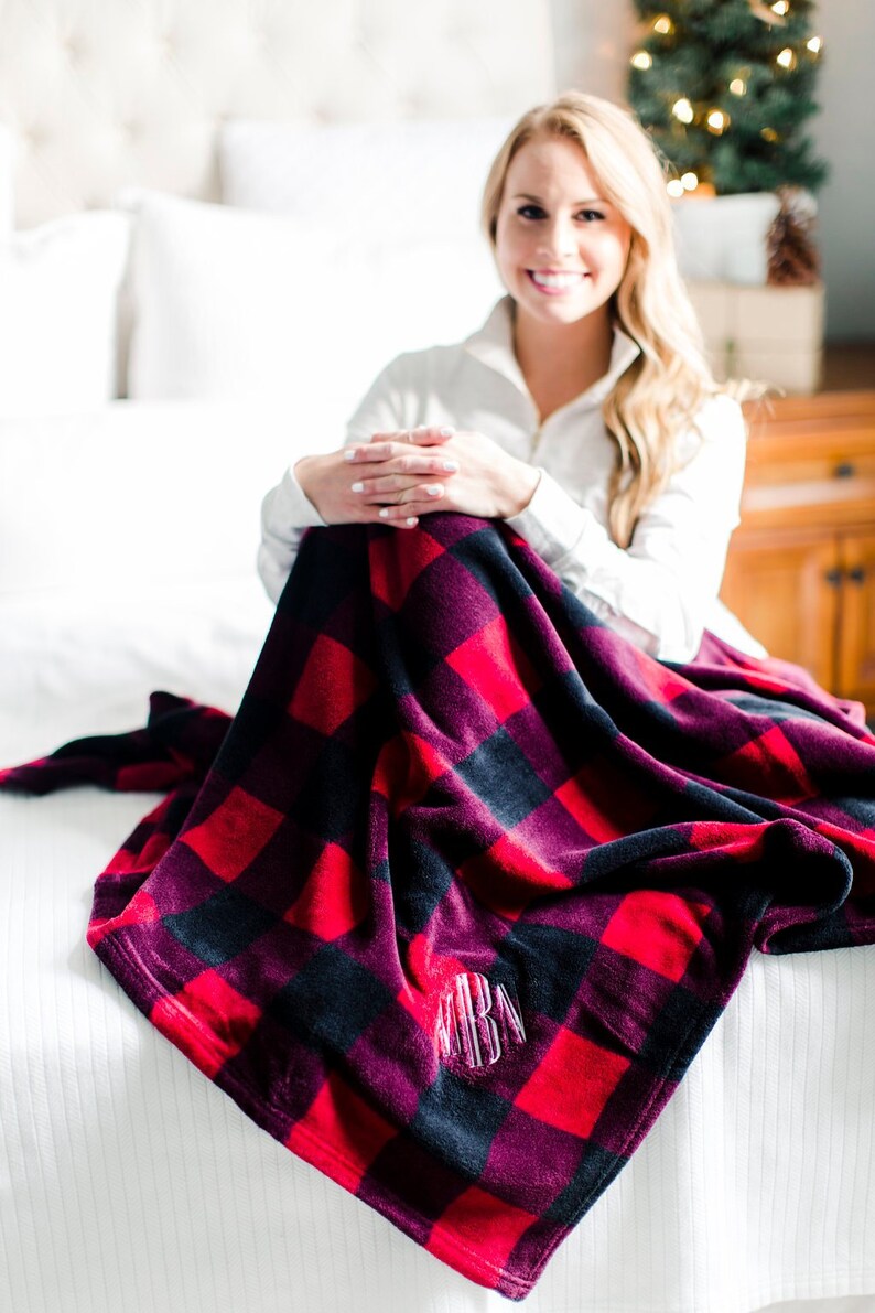 May include: A red and black plaid fleece blanket with a monogrammed initial in the center. The blanket is folded and draped over a white bed.