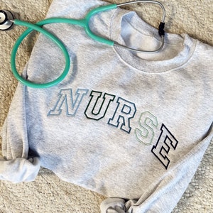 an ash crewneck sweatshirt with custom NURSE embroidered design in shades of blue and green