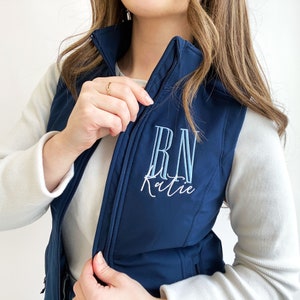 May include: A navy blue quilted vest with a zipper closure. The vest has "RN Katie" embroidered on the front in white and light blue thread.