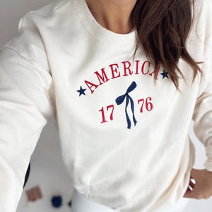 May include: Cream-colored sweatshirt with the word "AMERICA" arched across the chest in red embroidery, accompanied by blue stars and the number "1776". A blue bow graphic is centered below the word.