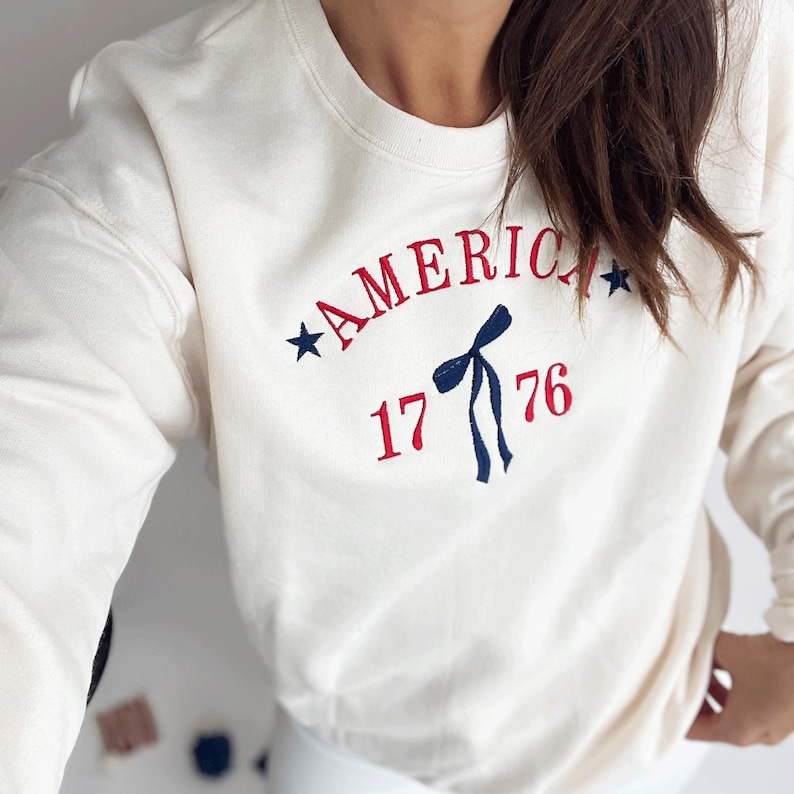 May include: Cream-colored sweatshirt with the word "AMERICA" arched across the chest in red embroidery, accompanied by blue stars and the number "1776". A blue bow graphic is centered below the word.