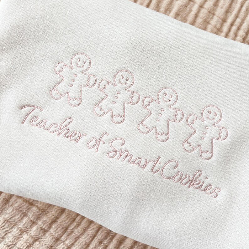 sweet cream crewneck sweatshirt with custom gingerbread men icons and "teacher of smart cookies" design embroidered in powder pink thread