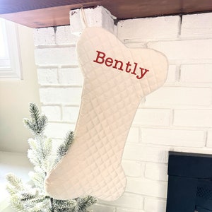 May include: Cream-colored, bone-shaped Christmas stocking with diamond quilting. The name "Bently" is embroidered in red. The stocking hangs from a white brick fireplace, with a small Christmas tree in the background.