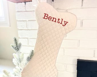 May include: Cream-colored, bone-shaped Christmas stocking with diamond quilting. The name "Bently" is embroidered in red. The stocking hangs from a white brick fireplace, with a small Christmas tree in the background.