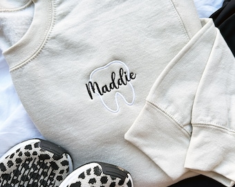 May include: A light beige sweatshirt with a white tooth graphic and the name "Maddie" embroidered in black.