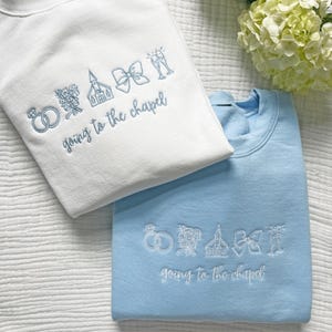 May include: Two crewneck sweatshirts, one white and one light blue, embroidered with wedding-themed icons and the phrase "going to the chapel" in a script font. The design includes rings, flowers, a church, a bow, and champagne flutes.