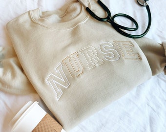 May include: Beige sweatshirt with the word "NURSE" embroidered in white and brown lettering.
