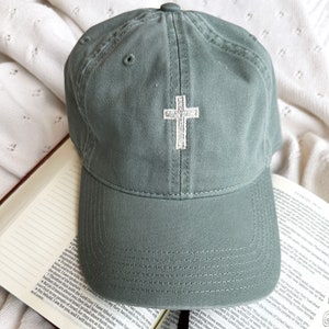May include: A green baseball cap with a white embroidered cross on the front. The cap is sitting on a white blanket and a book.
